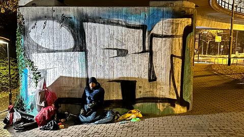 Der obdachlose Christian Endres sitzt in dicker Winterjacke, mit Mütze und Schlafsack unter einer Brücke, um sich vor Schneeregen zu schützen. 