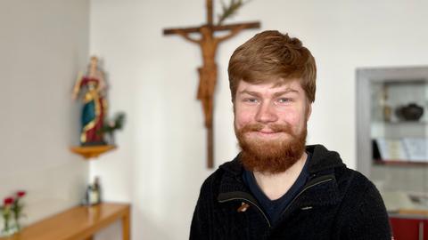 Peter Roberg steht in einem Zimmer mit religiösen Symbolen und lacht in die Kamera.