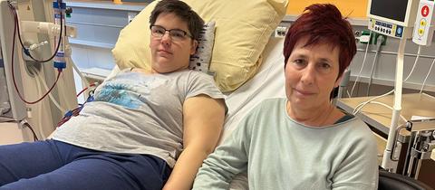 Saskia Wollenhaupt liegt auf Krankenhausbett, Mutter Sandra sitzt daneben, beide lächeln verhalten in die Kamera. 