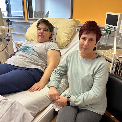 Saskia Wollenhaupt liegt auf Krankenhausbett, Mutter Sandra sitzt daneben, beide lächeln verhalten in die Kamera. 