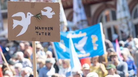 Im Vordergrund ein Schild mit Friedenstauben und den Worten "Waffen töten", im Hintergrund viele Menschen.