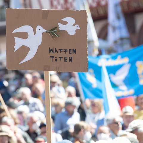 Im Vordergrund ein Schild mit Friedenstauben und den Worten "Waffen töten", im Hintergrund viele Menschen.