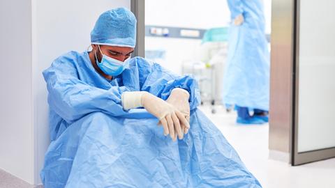 Erschöpfter Krankenhausmitarbeiter sitzt in Schutzkleidung auf dem Boden und starrt mit geneigtem Kopf vor sich hin.