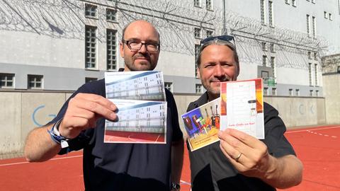 Zwei Männer vor einer Gefängnismauer halten Postkarten hoch