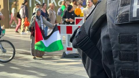 Das Bild zeigt am rechten Bildrand einen Polizisten in Uniform von hinten. Am linken oberen Bildrand sind Demonstranten hinter einer Absperrung zu sehen, eine Frau mit Kopftuch hält eine palästinensische Flagge.