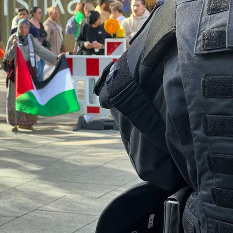 Das Bild zeigt am rechten Bildrand einen Polizisten in Uniform von hinten. Am linken oberen Bildrand sind Demonstranten hinter einer Absperrung zu sehen, eine Frau mit Kopftuch hält eine palästinensische Flagge.