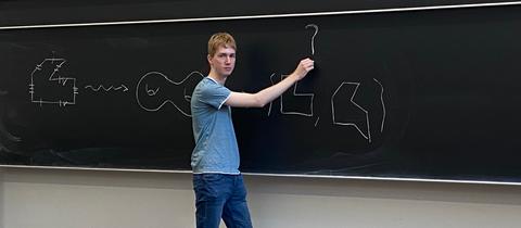 Ein junger Mann in Jeans und T-Shirt steht an einer großen Tafel in einem Hörsaal und zeichnet Formeln darauf .