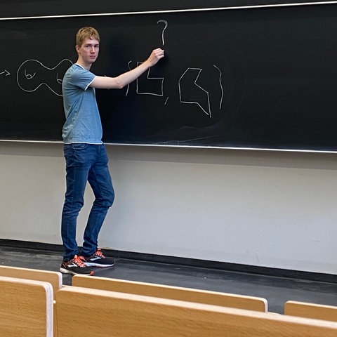 Ein junger Mann in Jeans und T-Shirt steht an einer großen Tafel in einem Hörsaal und zeichnet Formeln darauf .