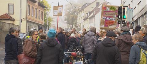 Dutzende Menschen stehen auf Straße und Bürgersteige bei einer Demo, einzelne Schilder werden in die Höhe gehalten.