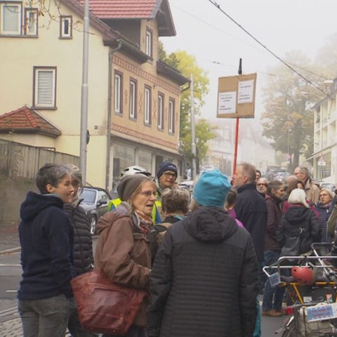 Dutzende Menschen stehen auf Straße und Bürgersteige bei einer Demo, einzelne Schilder werden in die Höhe gehalten.