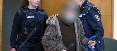 In einem Gerichtssaal wird eine Frau in einem grauen Mantel von zwei Justizwachtmeisterinnen auf die Anklagebank geführt.