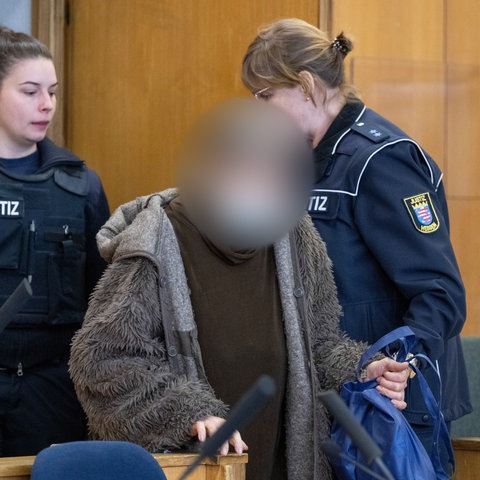 In einem Gerichtssaal wird eine Frau in einem grauen Mantel von zwei Justizwachtmeisterinnen auf die Anklagebank geführt.