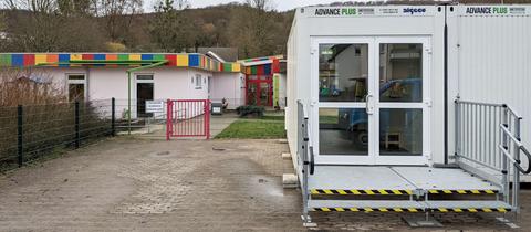 Weiße Container mit Eingangstür stehen neben einem Eingang zu einer Kita, in dessen Hof ein bunt angemaltes Gebäude steht.