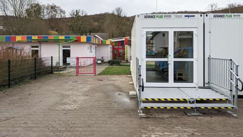 Weiße Container mit Eingangstür stehen neben einem Eingang zu einer Kita, in dessen Hof ein bunt angemaltes Gebäude steht.