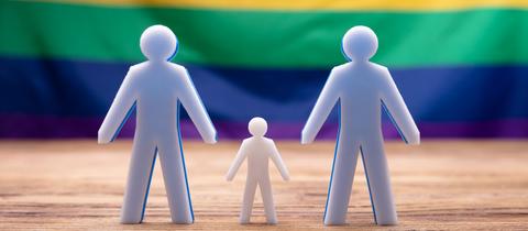 Figuren von zwei Männern und Kind vor Regenbogenfahne