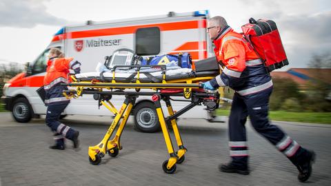 Zwei Rettungskräfte rennen mit einer Trage neben einem Einsatzfahrzeug mit der Aufschrift Malteser,