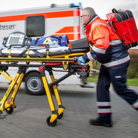 Zwei Rettungskräfte rennen mit einer Trage neben einem Einsatzfahrzeug mit der Aufschrift Malteser,