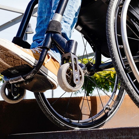 Rollstuhl mit Person in Nahaufnahme an einem Treppenabsatz - von unten fotografiert.