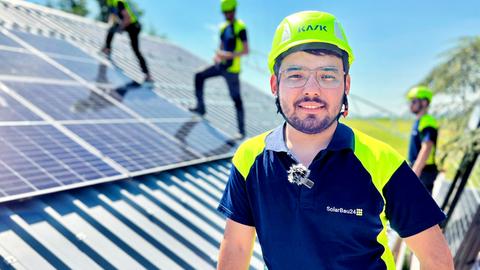 Im Bildvordergrund ein Mann mit Helm, der in die Kamera lächelt. Im Bildhintergrund unscharf drei Männer mit gelben Helmen auf einem Dach mit Solarpanelen.