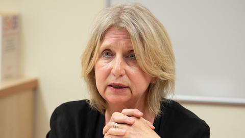 Eine blonde Frau in einem schwarzen Kostüm spricht mit ernstem Gesichtsausdruck.