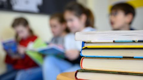 Bücher liegen auf einem Schreibtisch in einem Klassenraum, im Hingergrund Kinder