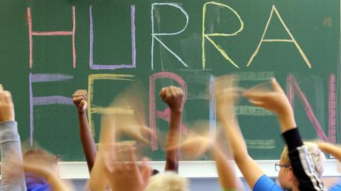 Schultafel mit der Aufschrift "Hurra Ferien" - im Vordergrund gereckte Kinderhände