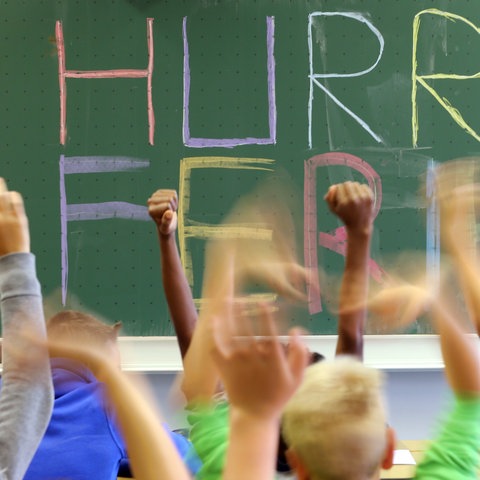 Schultafel mit der Aufschrift "Hurra Ferien" - im Vordergrund gereckte Kinderhände