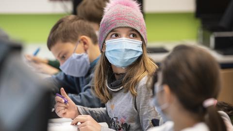 Mit Mund-Nase-Masken sitzen Schülerinnen und Schüler im Unterricht. Ein Mädchen schaut auf und sieht in Richtung Kamera.