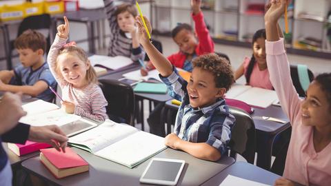 Schülerinnen und Schüler sitzen in Reihen vor einer Lehrkraft, melden sich und lächeln.