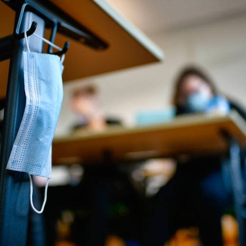 Eine Maske hängt an einem Tisch im Klassenzimmer am Haken, an dem früher die Schultaschen hingen. Im Hintergrund sind unscharf Schüler mit Masken an Tischen sitzend zu sehen.