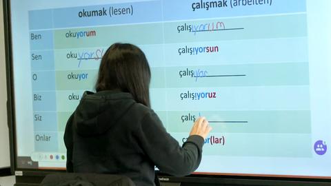 Eine Person (von hinten fotografiert) steht vor einem Smartboard, auf dem Verben der türkischen Sprache in einer Tabelle angeordnet sind. 