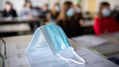 Eine OP-Maske liegt auf einem Schultisch in einem Klassenzimmer. ImHintergrund sind Schülerinnen und Schüler leicht verschwommen zu sehen.