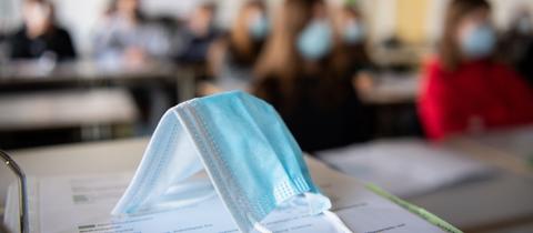 Eine OP-Maske liegt auf einem Schultisch in einem Klassenzimmer. Im Hintergrund sind Schülerinnen und Schüler leicht verschwommen zu sehen.