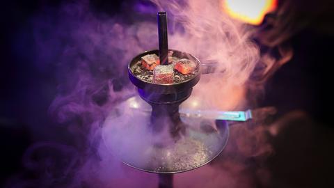 Eine Shisha Wasserpfeife mit drei glühenden Kohlen auf dem Kaminaufsatz in einer Shisha-Bar. Rauch und Neonlichter im Hintergrund.
