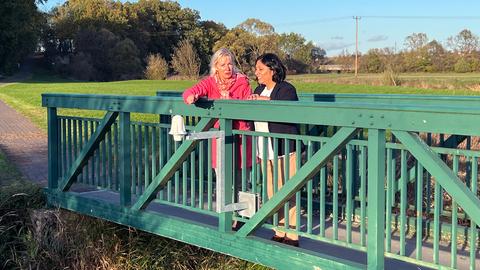 Kristina Sinemus und Ramona-Margarita Ruppert stehen in der Abendsonne auf einer Fußgängerbrücke, an welcher ein Senor angebracht ist.