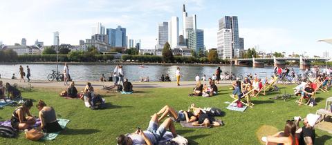 Personen genießen die warme Sonne und den Spätsommertag auf der Liegewiese am Mainufer vor dem Bankenviertel in Frankfurt