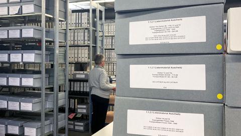 Blick in das Archiv der Arolsen Archives: Im Vordergrund liegen graue Kartons mit Unterlagen, fein säuberlich beschriftet. Im Hintergrund sieht man eine grauhaarige Frau zwischen hohen Regalen.