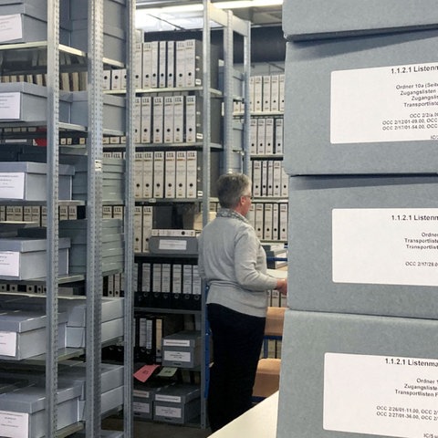 Blick in das Archiv der Arolsen Archives: Im Vordergrund liegen graue Kartons mit Unterlagen, fein säuberlich beschriftet. Im Hintergrund sieht man eine grauhaarige Frau zwischen hohen Regalen.