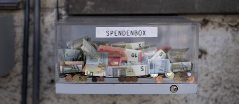 Transparente Box mit Geldmünzen und Geldscheinen - mit der Aufschrift "Spendenbox"