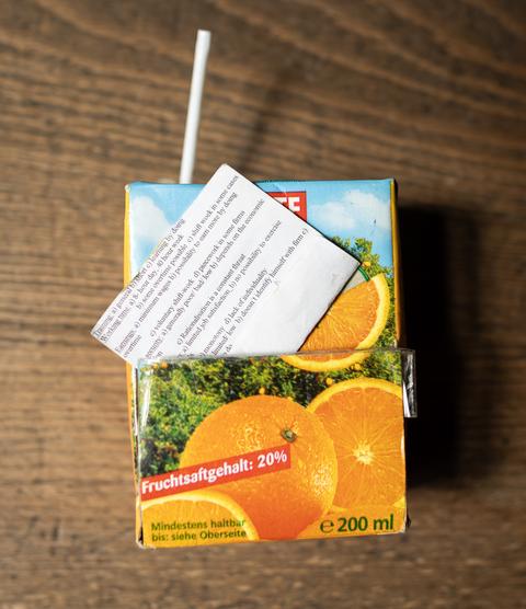 Ein Trinkpäckchen mit Orangensaft. Aus einem Schlitz in der Mitte ragt ein kleiner Zettel mit kleingedruckter Schrift heraus.