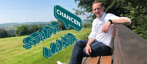 Der junge Bürgermeister von Feldatal sitzt auf einer überdimensionalen Holzbank, die mitten in der Landschaft steht (von der Seite fotografiert). Daneben das Logo der ARD-Studie "Stadt Land Chancen".