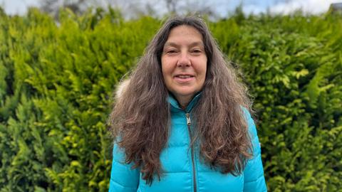 Frau mit langen braunen Locken steht vor einer grünen Hecke im Garten