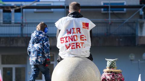 Rückansicht eines Mannes, der auf einem kugelförmigen Stein sitzt und eine weiße Plastikweste trägt, auf welcher in roten Buchstaben "Wir sind es wert" steht. Daneben sitzen zwei Kinder.