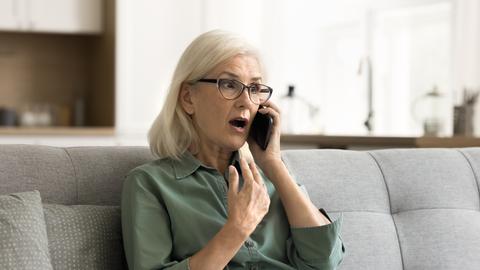 Eine ältere Frau sitzt auf einem grauen Sofa und bekommt gerade per Telefon eine schockierende Nachricht