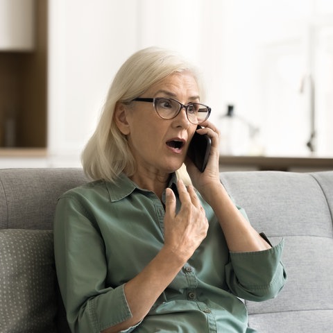 Eine ältere Frau sitzt auf einem grauen Sofa und bekommt gerade per Telefon eine schockierende Nachricht