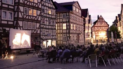 Vor einem Fachwerkhaus ist eine Leinwand aufgebaut. Davor sitzen viele Menschen auf Stühlen und schauen einen Film.