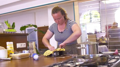 Eine Frau mit weiß-blau gestreiftem T-Shirt und blonden, zum Zopf gebundenen Locken steht in einer Küche und gibt Teig in eine Springform.