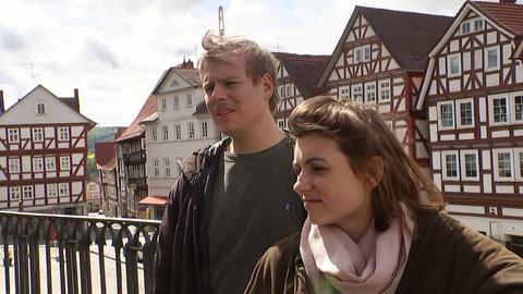 Ein Mann und eine Frau stehen auf einer Terrasse. Hinter ihnen sind Fachwerkhäuser zu sehen.