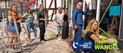 Männer und Frauen stehen vor einem leeren Ladengeschäft. Mit buntem Klebeband bekleben sie ein Schaufenster. 