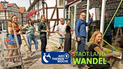 Männer und Frauen stehen vor einem leeren Ladengeschäft. Mit buntem Klebeband bekleben sie ein Schaufenster. 
